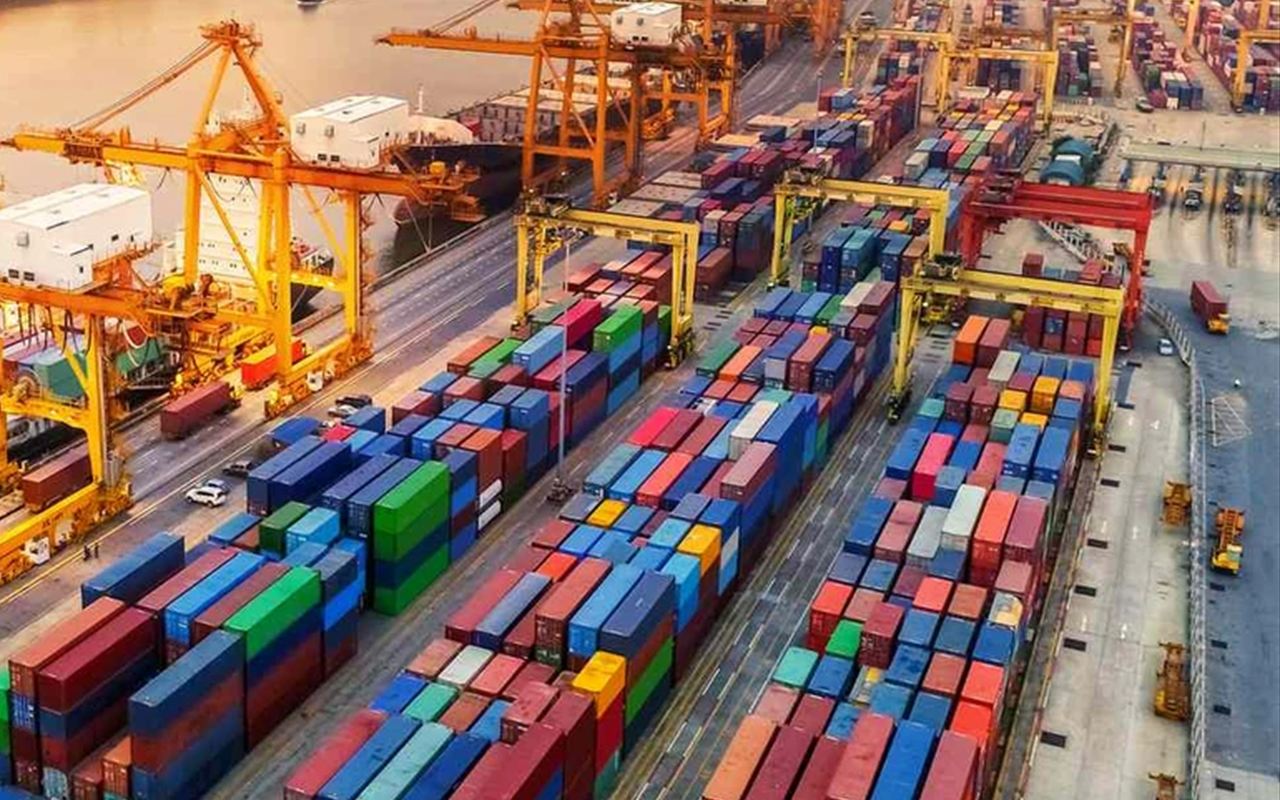 Rows of shipping containers stacked in a port yard beside gantry cranes.