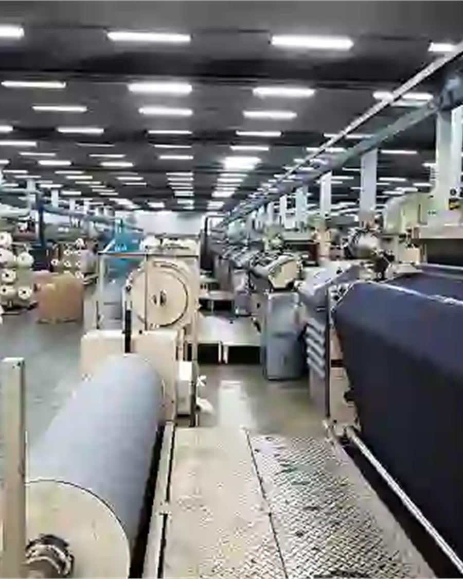 Rows of industrial looms and fabric rolls inside a textile weaving mill.